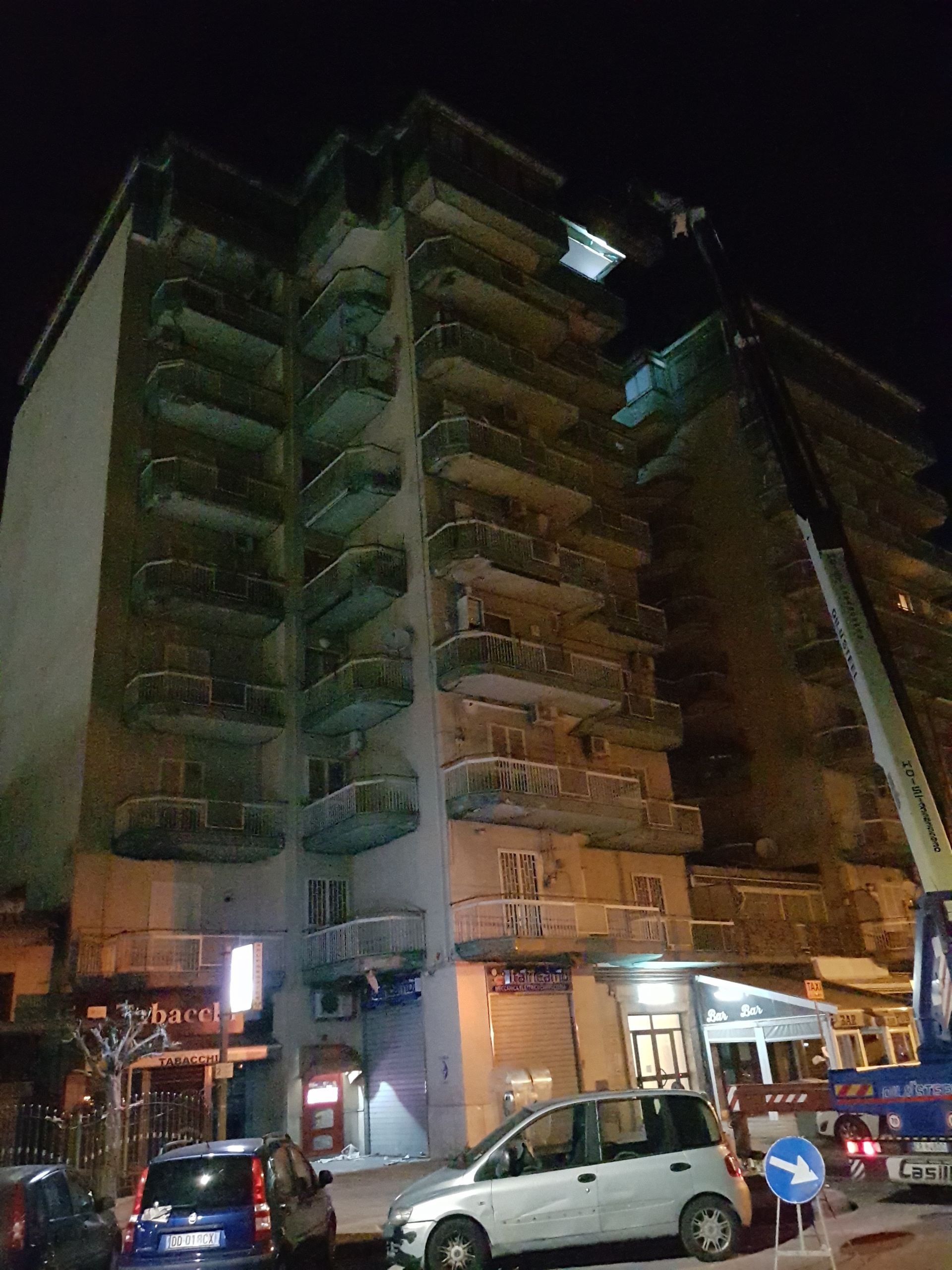 Veduta notturna di un alto condominio con balconi illuminati e auto parcheggiate al piano inferiore.
