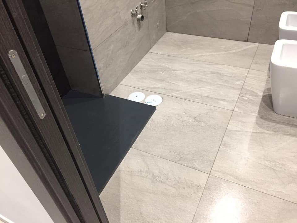 Ingresso del bagno con pavimento in piastrelle beige, tappetino scuro e sanitari visibili sulla destra.