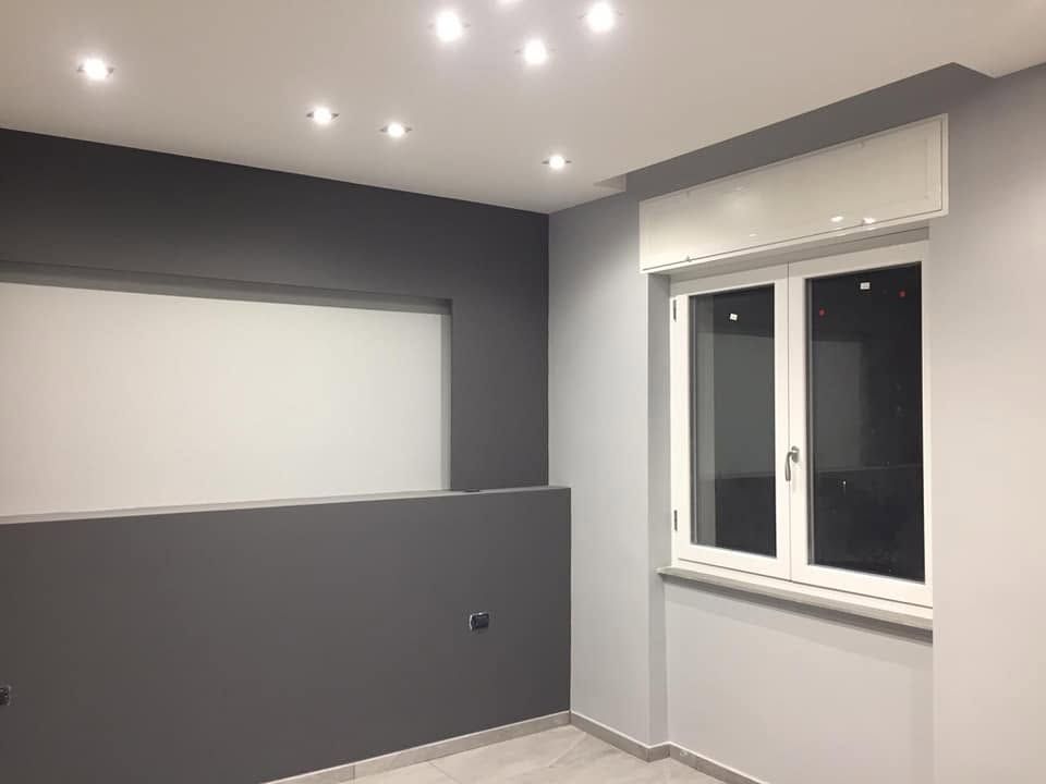 Camera moderna con pareti grigie, faretti a incasso sul soffitto, una tenda bianca e un'ampia finestra.