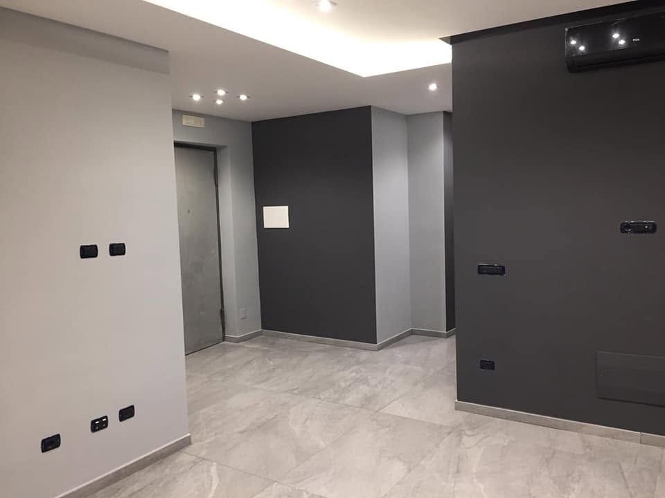 Stanza moderna vuota con pareti grigie, pavimento in piastrelle chiare, faretti a incasso sul soffitto e una porta sul retro.