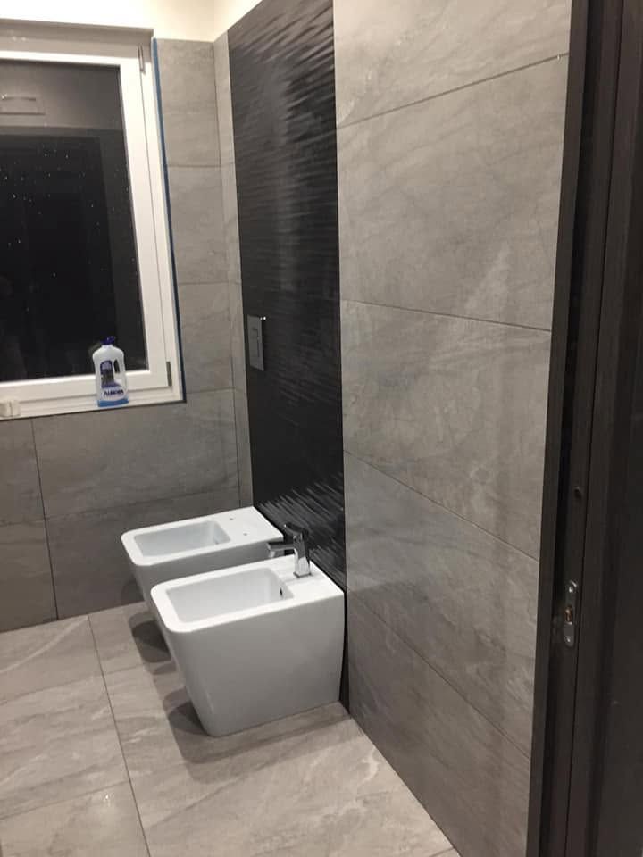 Bagno moderno piastrellato con finestra, bidet e WC accanto a una parete con dettagli neri.