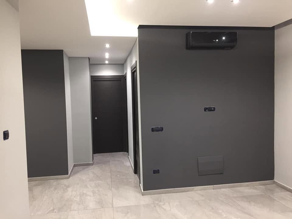 Interno di un appartamento moderno vuoto con pareti grigie, pavimento in piastrelle bianche e corridoio che conduce a una porta scura.