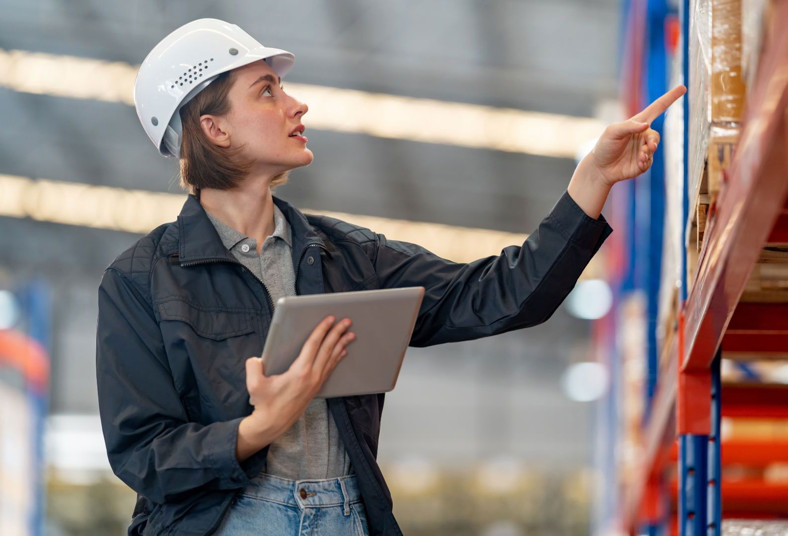 A woman is holding a tablet and pointing at a shelf in a warehouse.