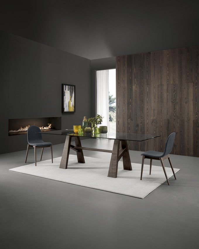 In una sala da pranzo moderna e minimalista, un tavolo da pranzo con piano in vetro e gambe in legno scuro, abbinato a due sedie grigie, è posizionato su un tappeto bianco.