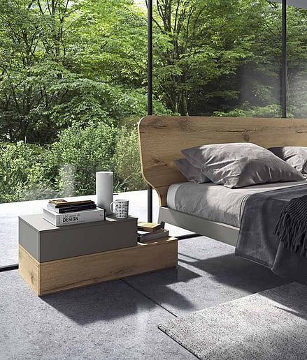 Una camera da letto minimalista con letto in legno, comodino grigio e un'ampia finestra a tutta altezza con vista sulla natura.