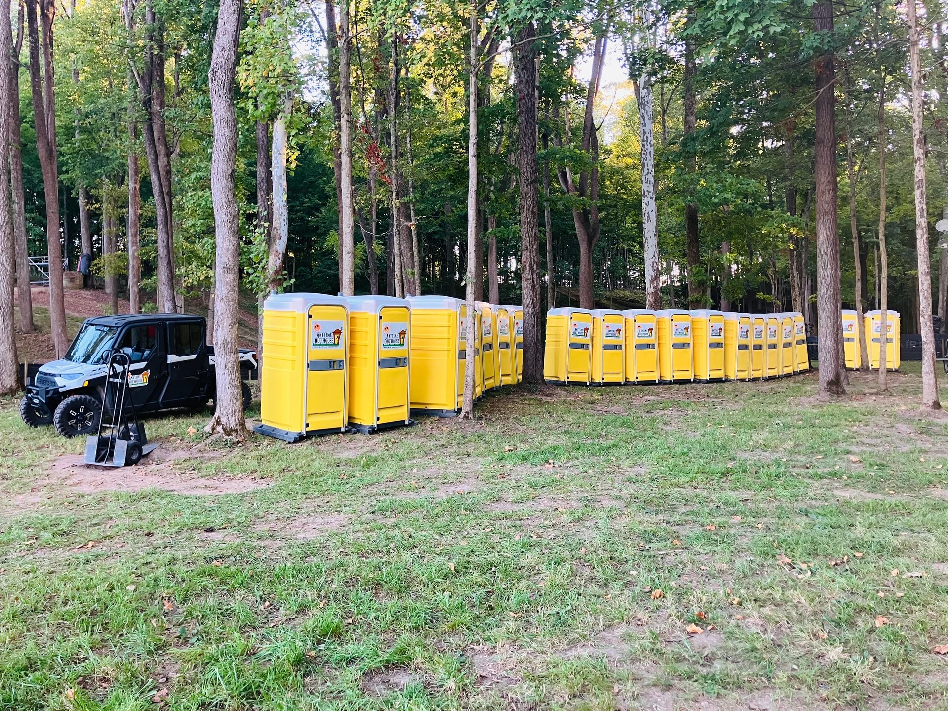 Yellow portable toilets lined up in a grassy area next to trees with a black vehicle.