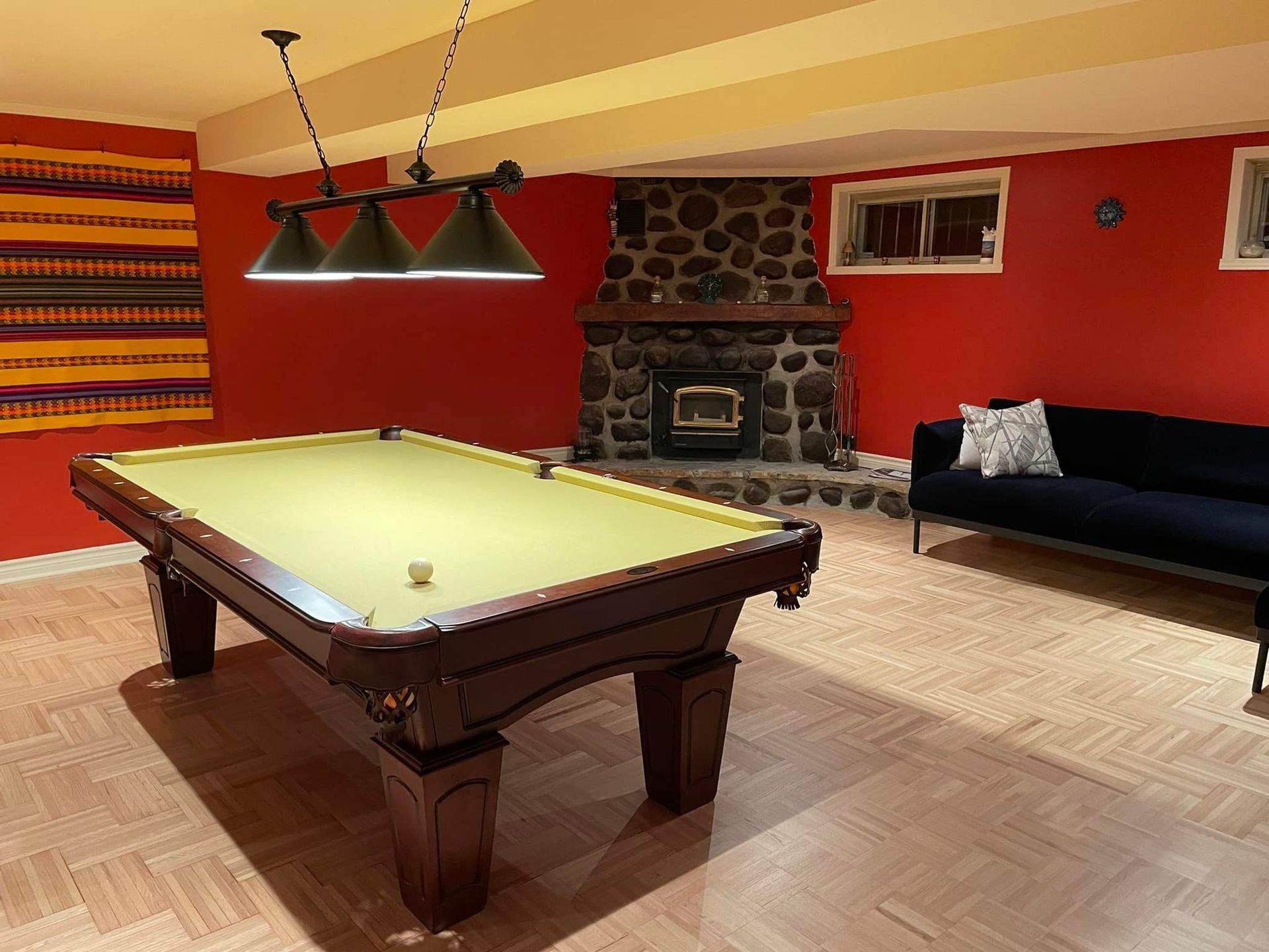 Une table de billard dans une pièce avec une cheminée en pierre, des murs rouges et un canapé noir.