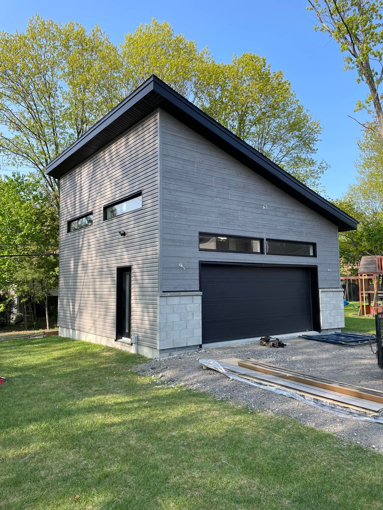 Garage gris moderne avec porte et toit noirs, petites fenêtres. Allée gravillonnée, pelouse verte, arbres.