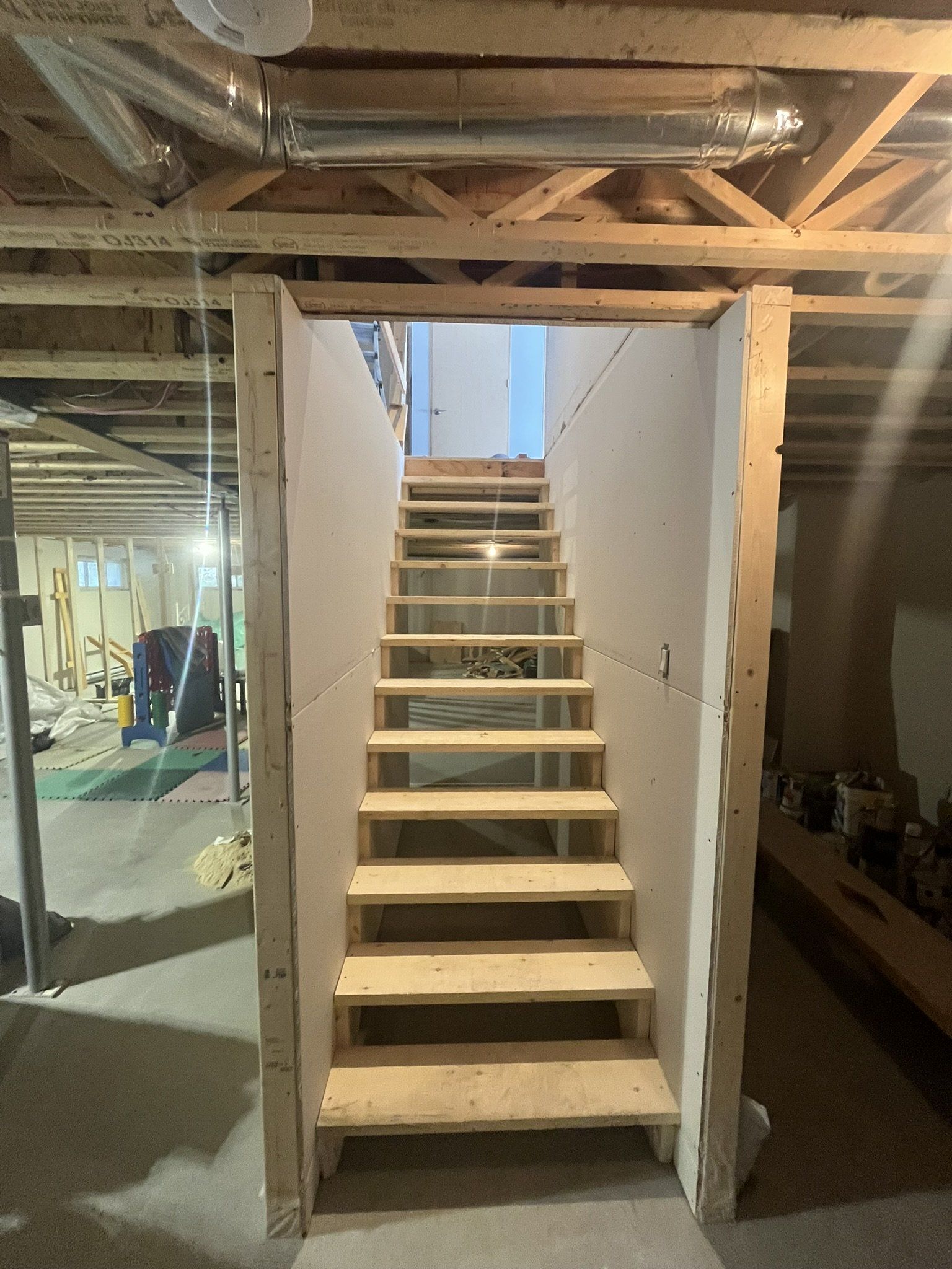Escalier en bois montant vers l'étage, encadré par des murs inachevés, dans un sous-sol en construction.
