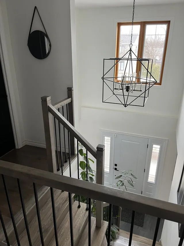 Vue depuis le palier d'un escalier. Escalier avec rampe en bois et balustres noirs, menant à une porte d'entrée avec imposte. Luminaire décoratif géométrique.