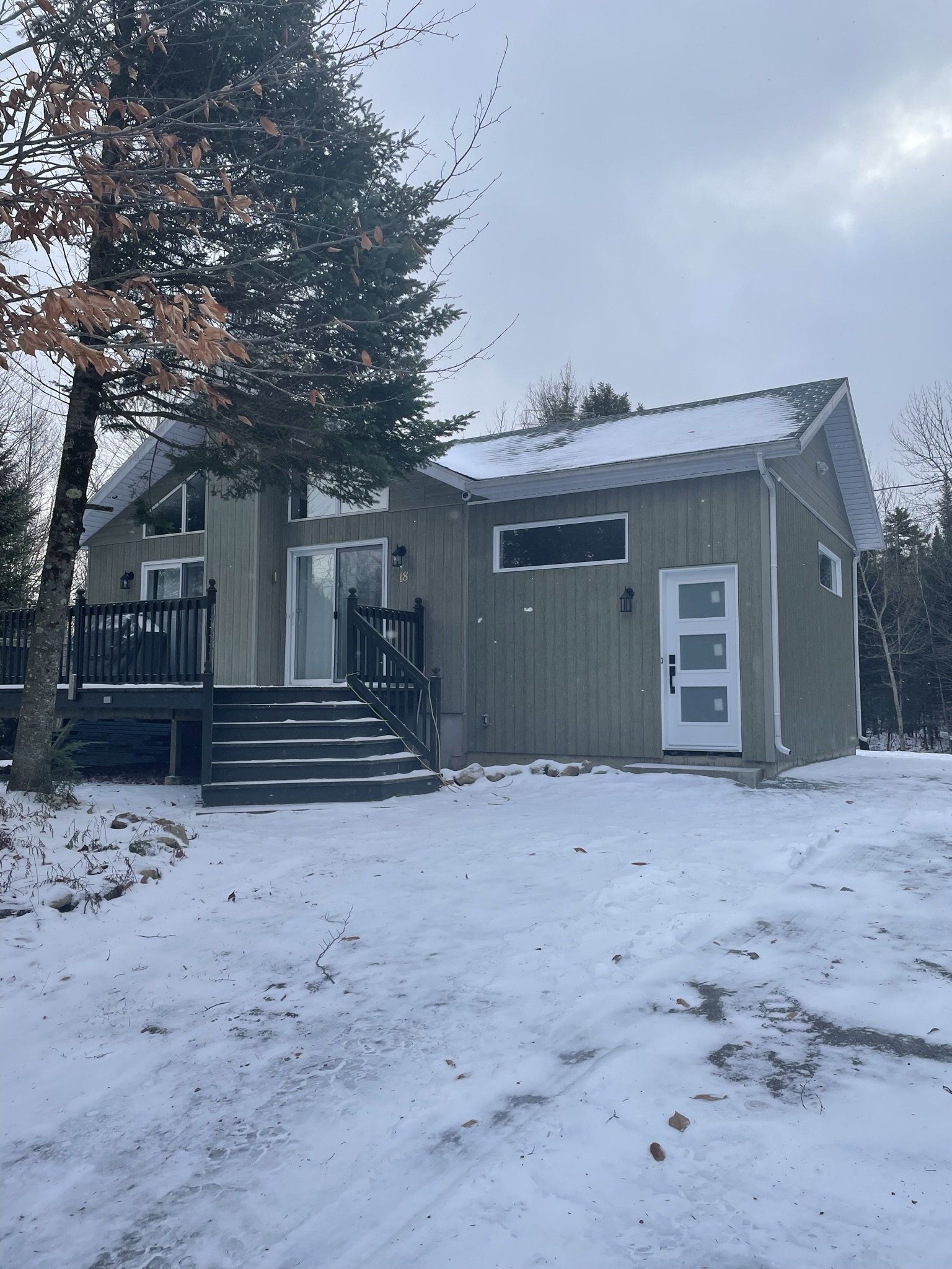 Extérieur d'un chalet sous la neige, avec terrasse et porte d'entrée. Bardage gris, porte blanche et sol enneigé.
