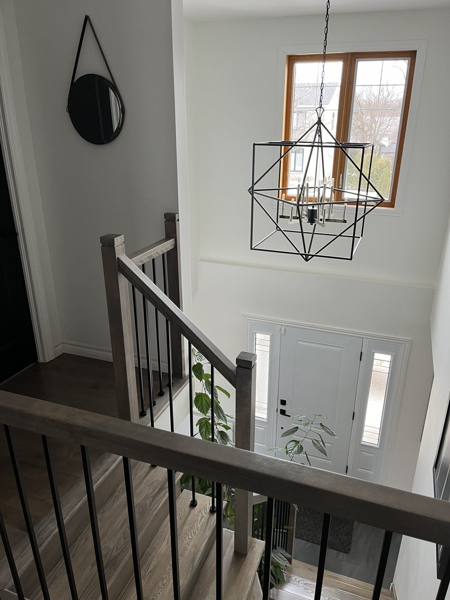 Vue depuis le palier d'un escalier. Escalier avec rampe en bois et balustres noirs, menant à une porte d'entrée avec imposte. Luminaire décoratif géométrique.