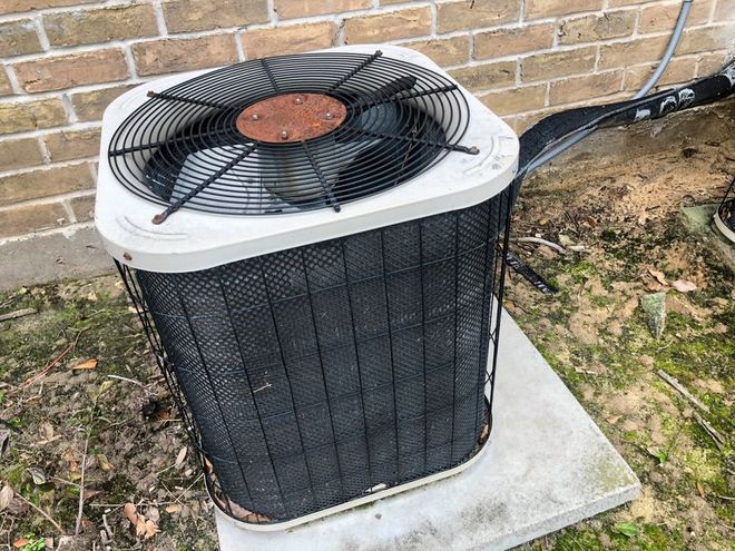 Outdoor air conditioner unit with black fan grille on a concrete pad beside a brick wall.
