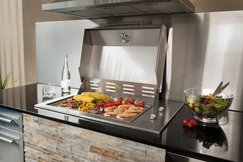 Modern outdoor grill built into a black countertop with food cooking inside a stainless steel hood.