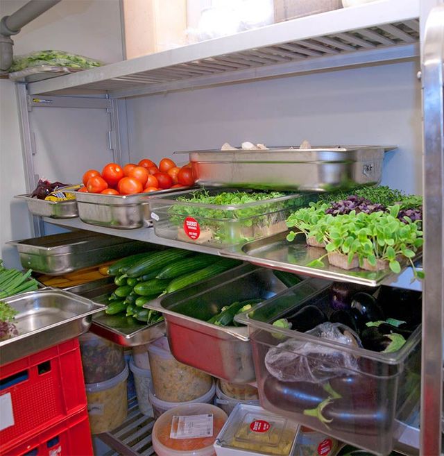 Shelves of fresh produce in a refrigerated prep station, including tomatoes, cucumbers, greens, and herbs.