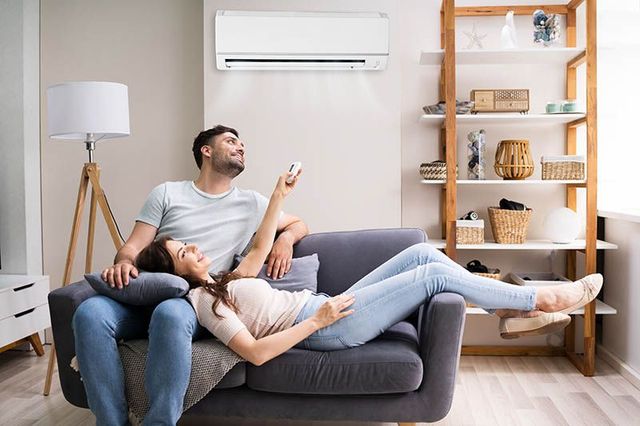 Couple relaxing on a gray sofa in a bright living room, with a wall-mounted air conditioner and wooden shelf.