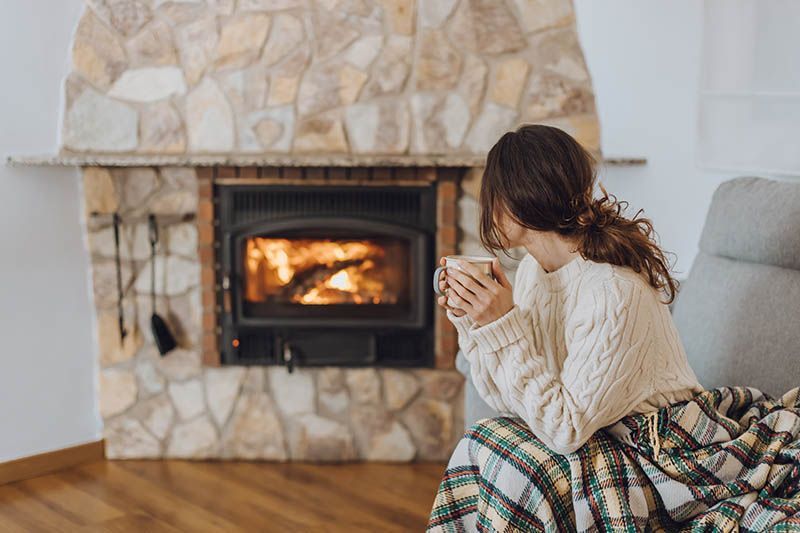 Person in a sweater sits by a lit stone fireplace, holding a mug and wrapped in a plaid blanket.