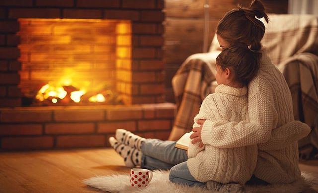 Child in a cream sweater sits on the floor by a glowing fireplace, hugging a stuffed toy with a mug nearby.