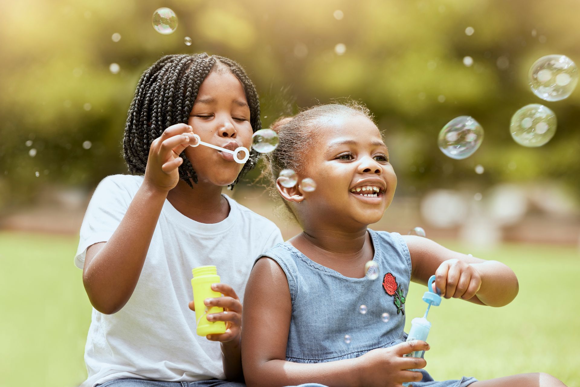 Two young girls are in the park and blowing bubbles. They are smiling.