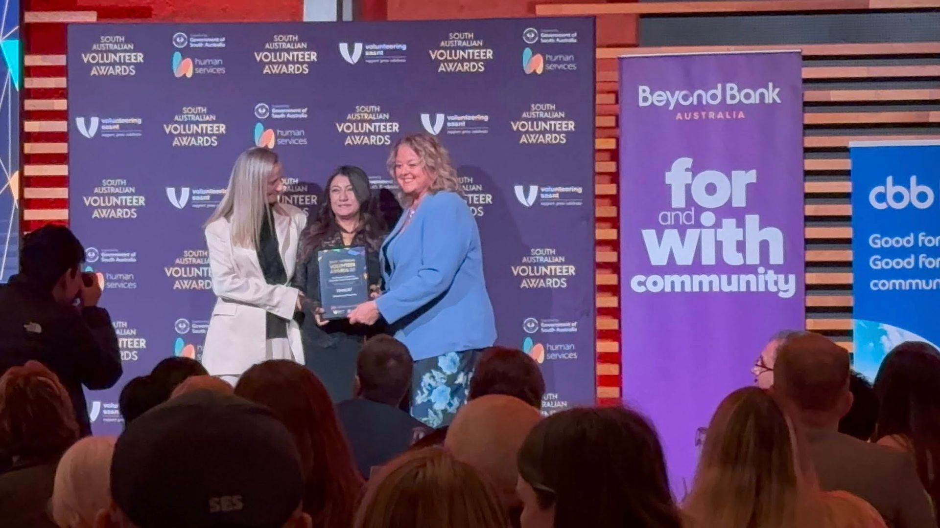 Three people holding an award onstage at a community event with sponsor logos behind them