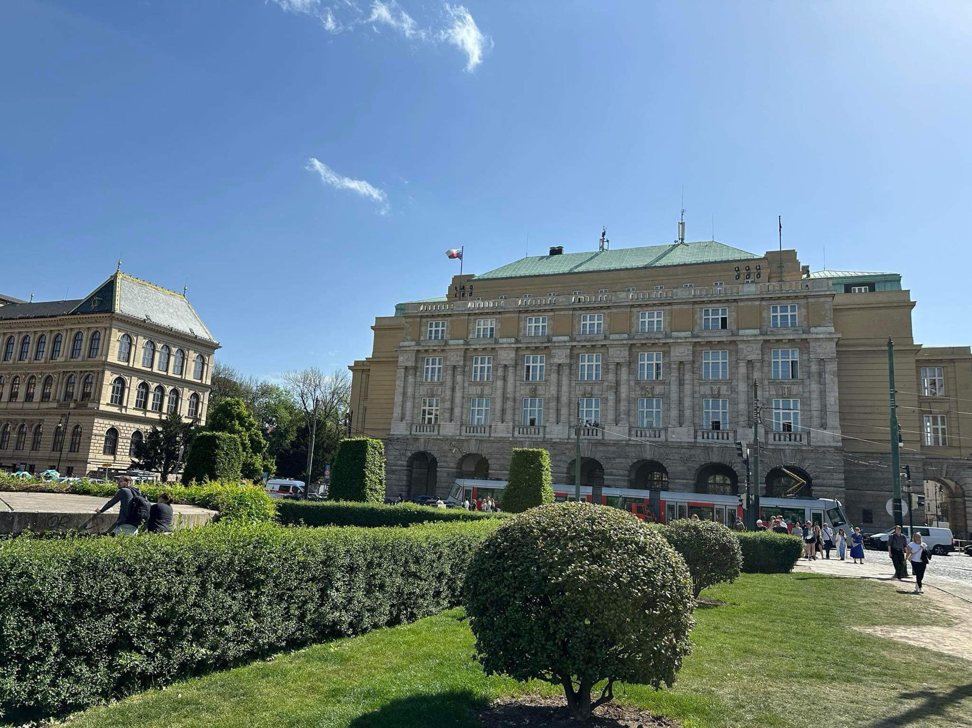 Sunny city square with manicured hedges and historic stone buildings under a blue sky