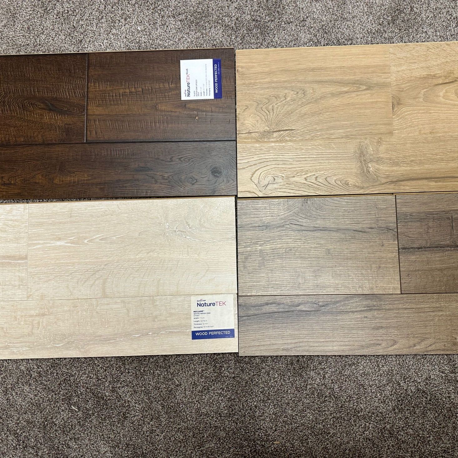 Four wood flooring samples, varying shades of brown and beige, arranged on a gray carpet.
