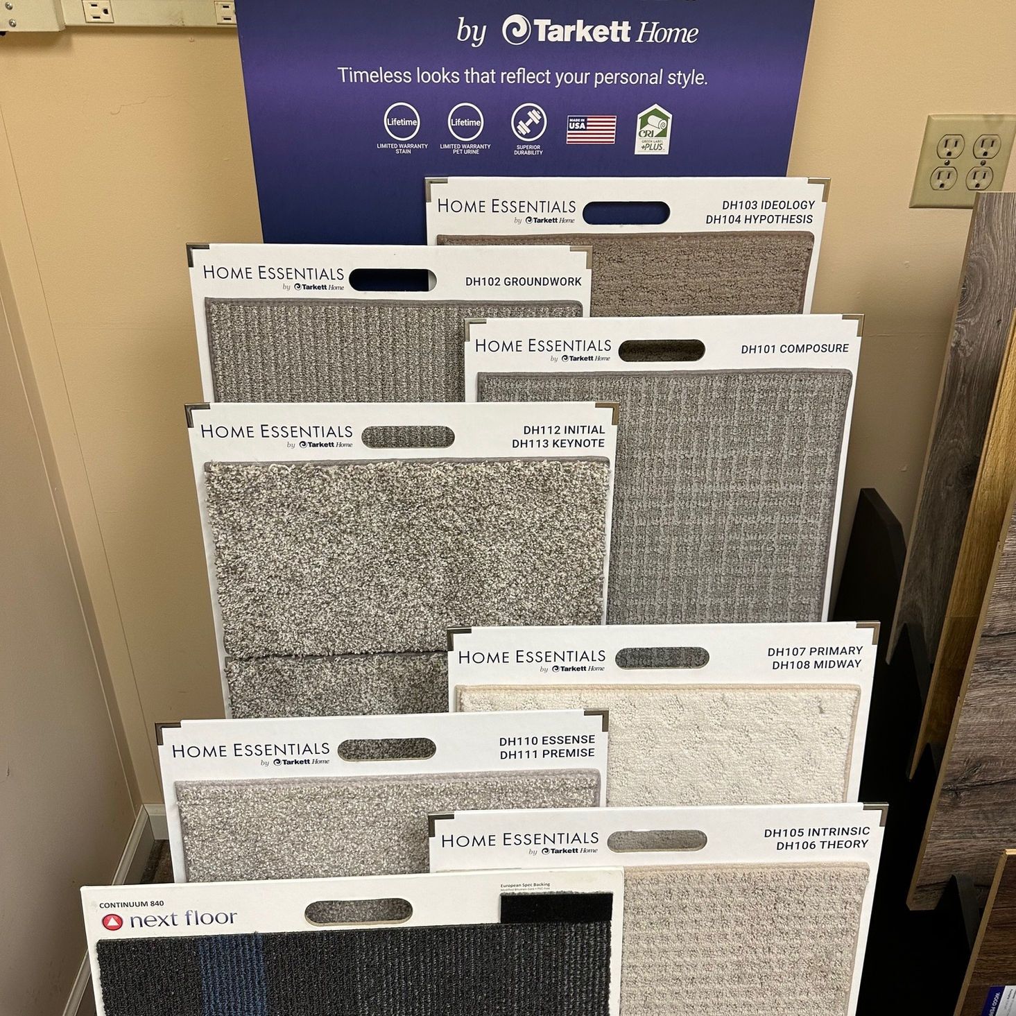 Carpet samples displayed on a rack. Gray and beige tones dominate. Marketing sign in the background.