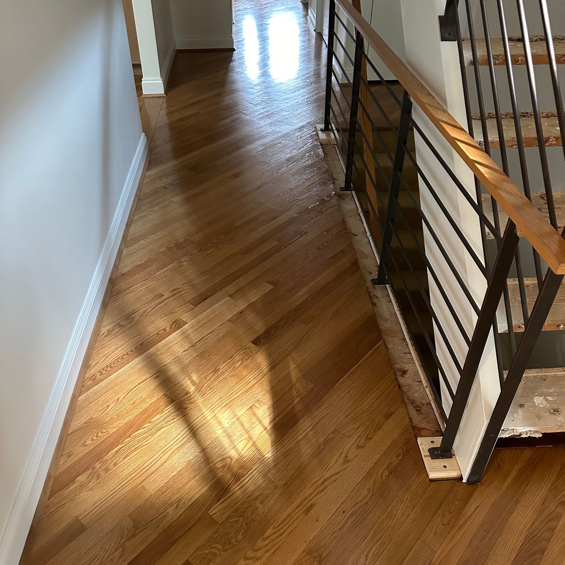 Herringbone patterned wood flooring in hallway, next to staircase with black railing and wood handrail.