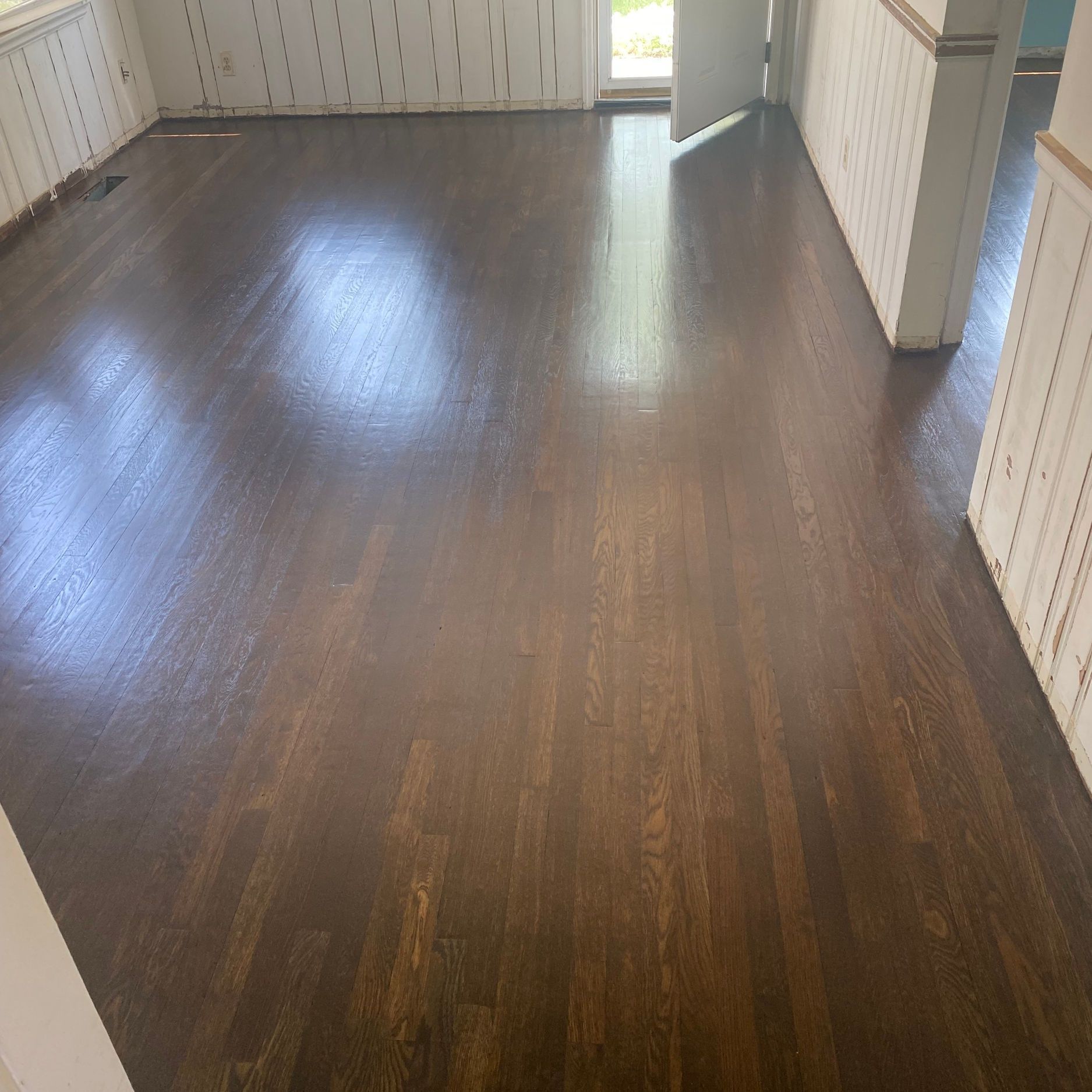 Dark stained wooden floor in a room with white paneled walls and a doorway to outside.