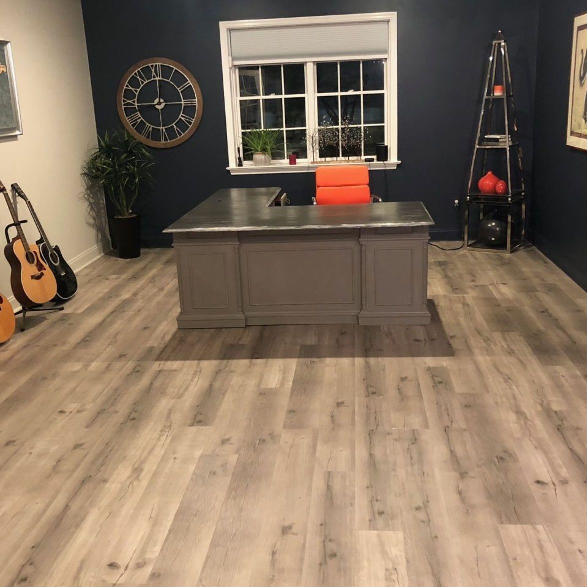 Home office with gray desk, guitars, clock, and orange chair, with wood-look flooring.