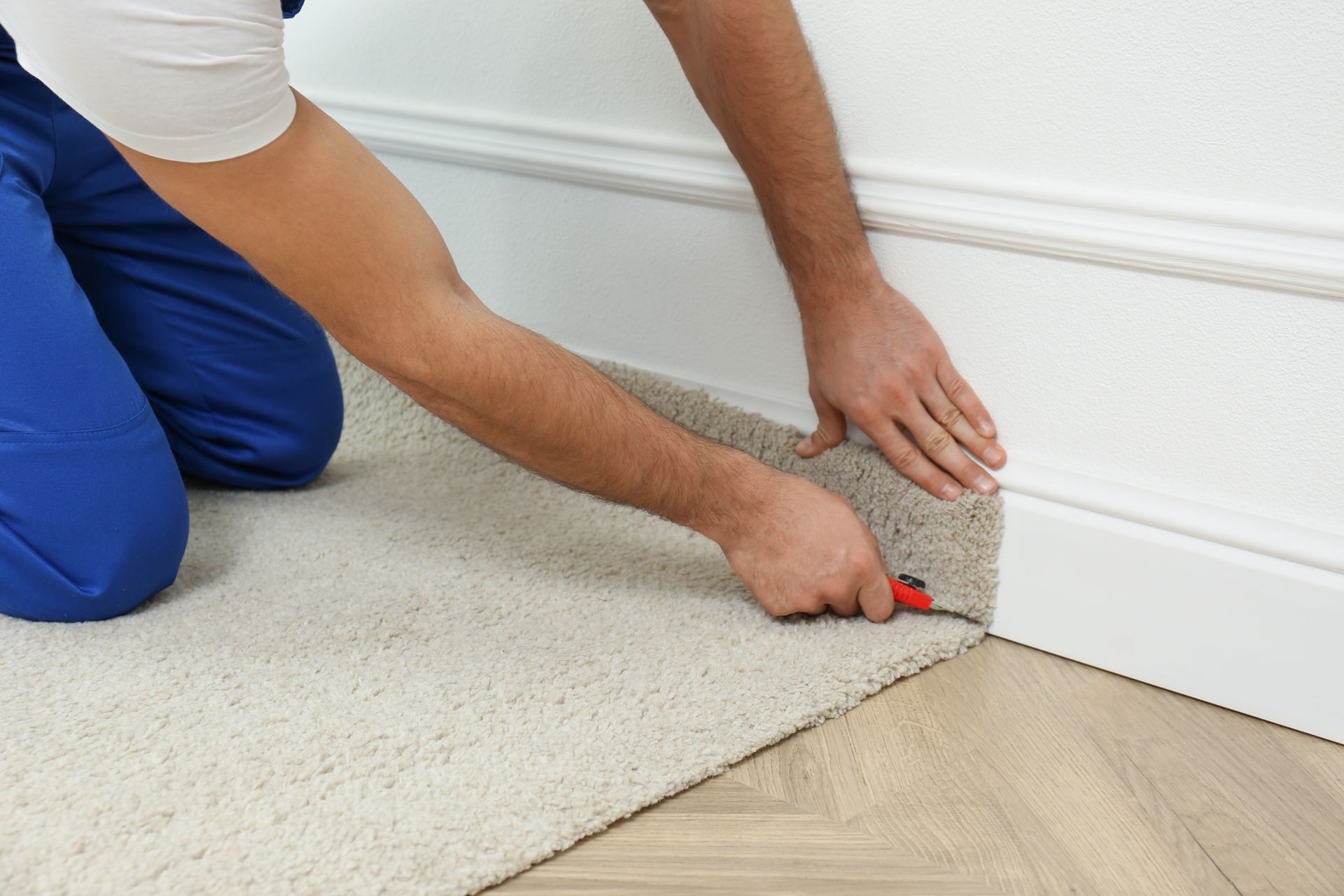 Person using a utility knife to trim carpet along a baseboard in a room.