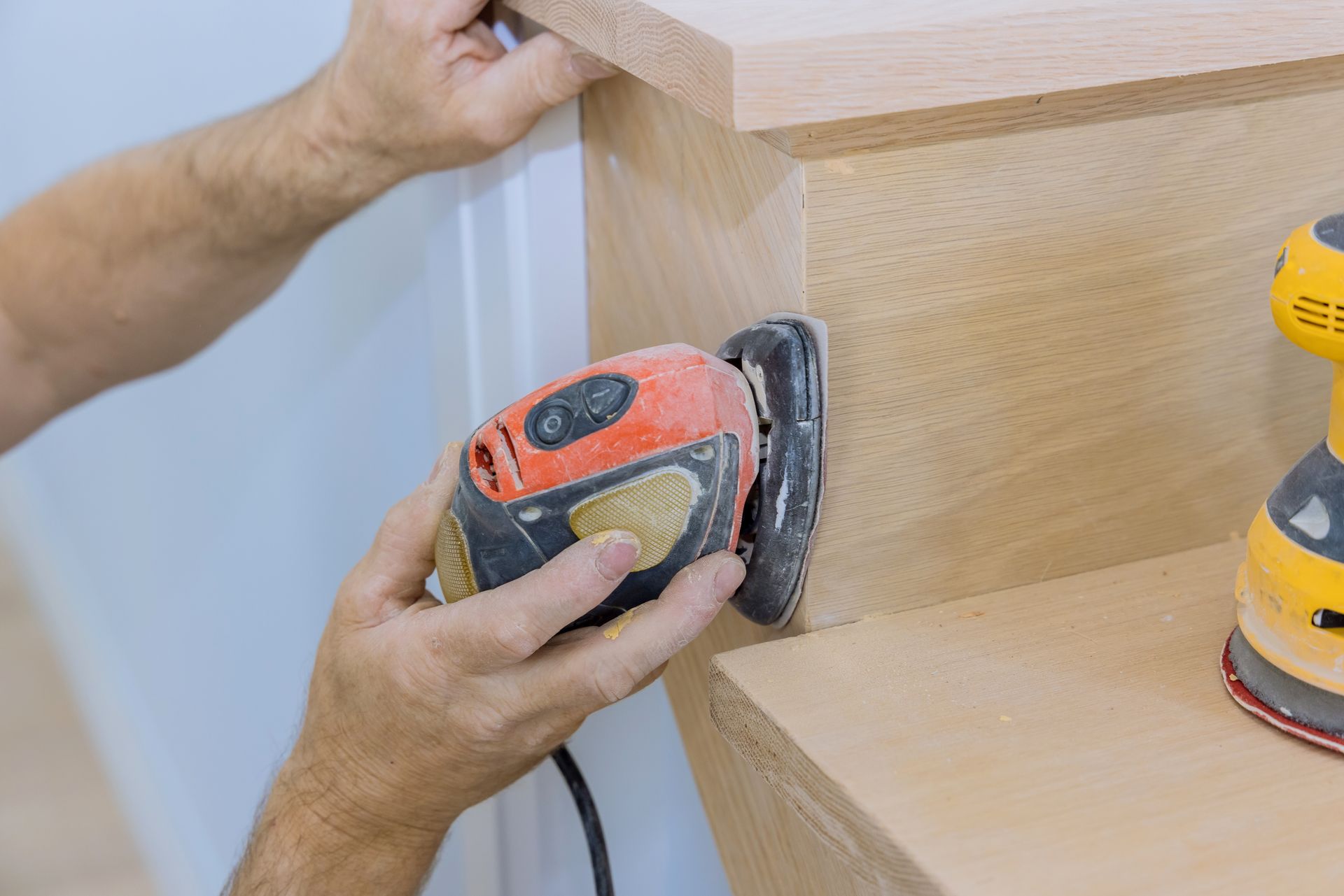 A person uses an orange handheld sander to smooth the wooden surface of a staircase.