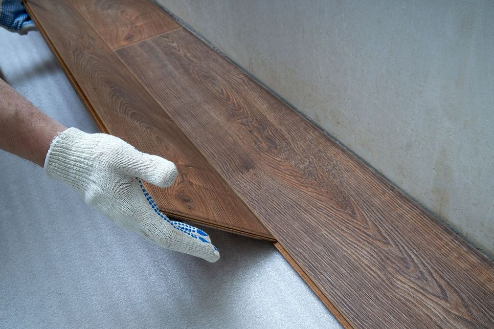 Gloved hands installing a brown wood plank flooring panel over white underlayment