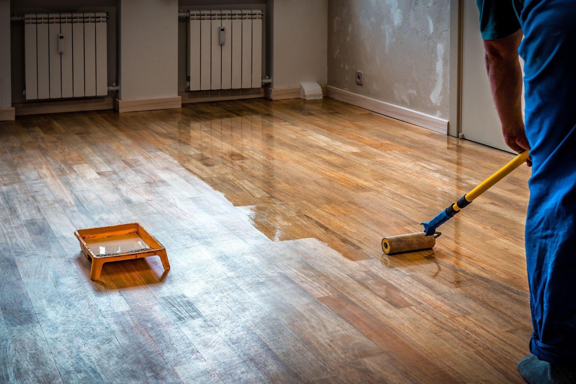 A person in blue overalls applies varnish to a wooden floor with a paint roller in a room with two wall heaters.