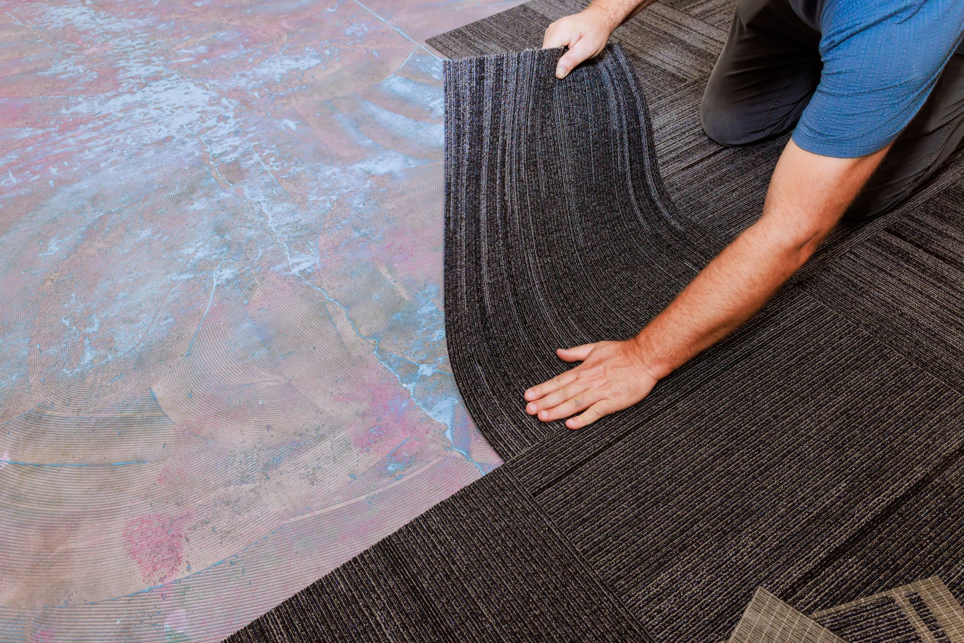 Person rolling out a dark textured carpet on a floor with a blue and brown surface.