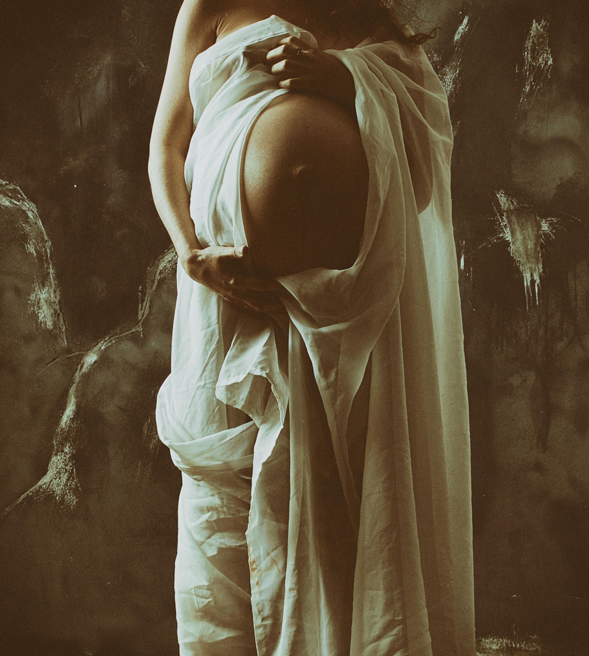 Pregnant person holding belly, wrapped in white fabric. Dark background, sepia tones.