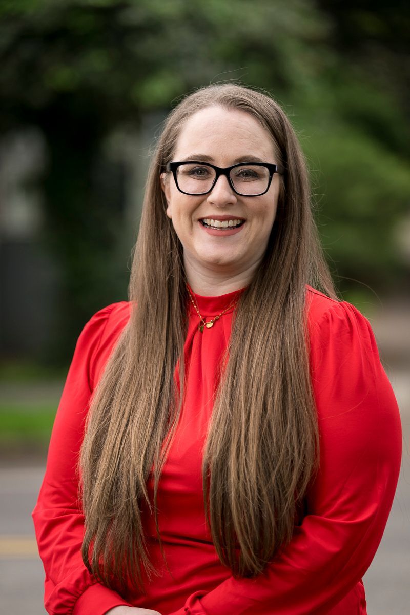 Woman with long brown hair wearing glasses and a red dress, smiling in front of a blurred outdoor background.