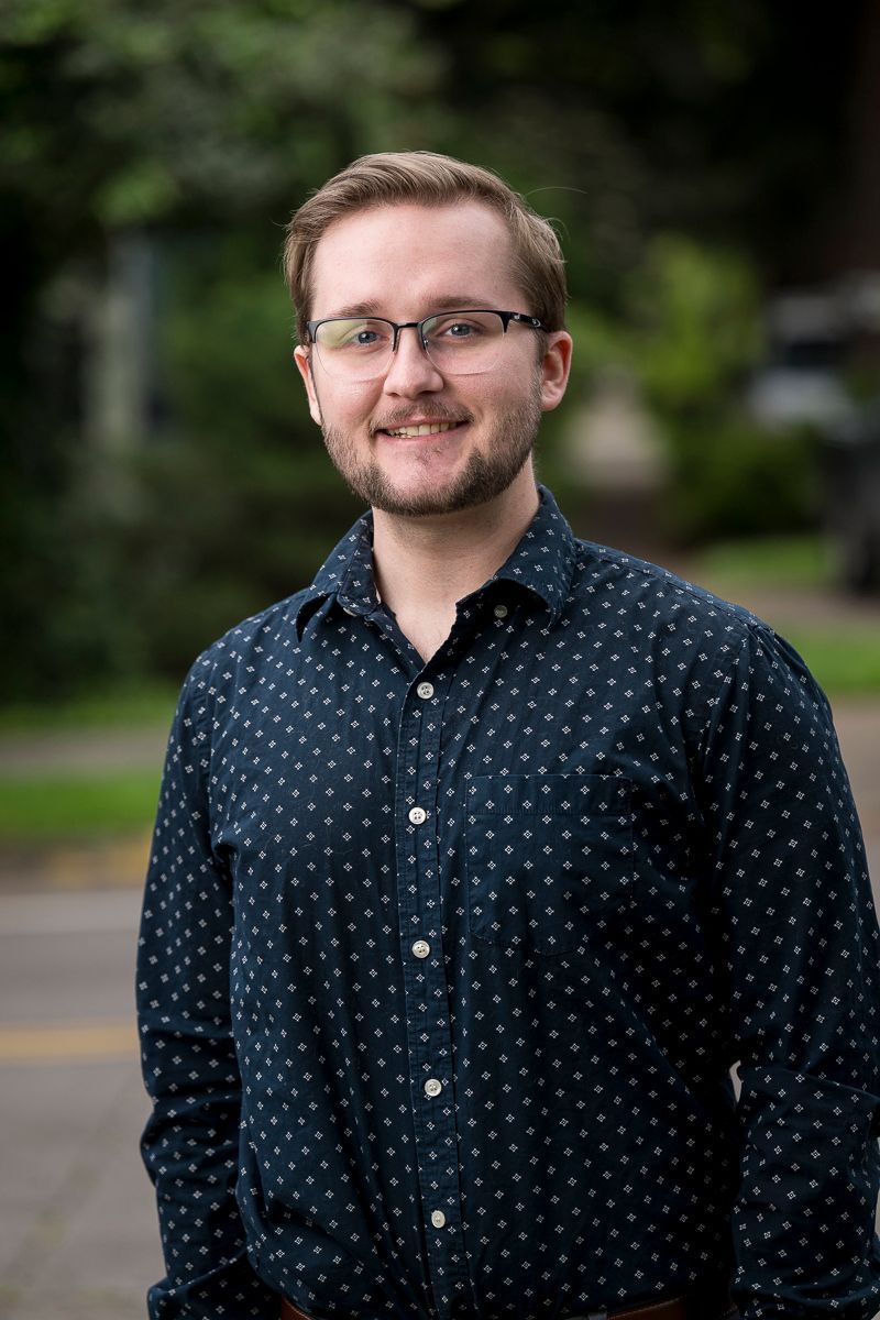 Man with glasses and short blonde hair, smiles slightly, wearing navy buttoned shirt, with blurred outdoor background.