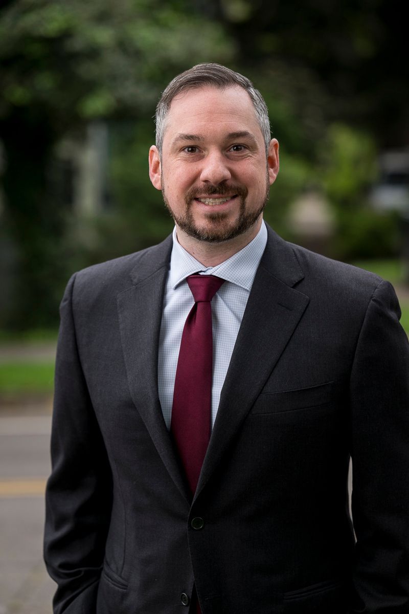 Man in suit smiles at the camera, dark hair and beard, red tie, blurred outdoor background.