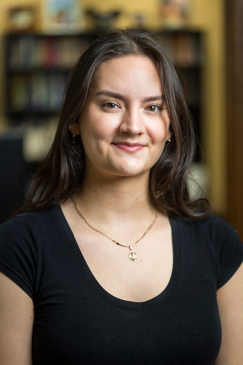 Woman smiling, wearing black top and gold necklace, in a library setting.