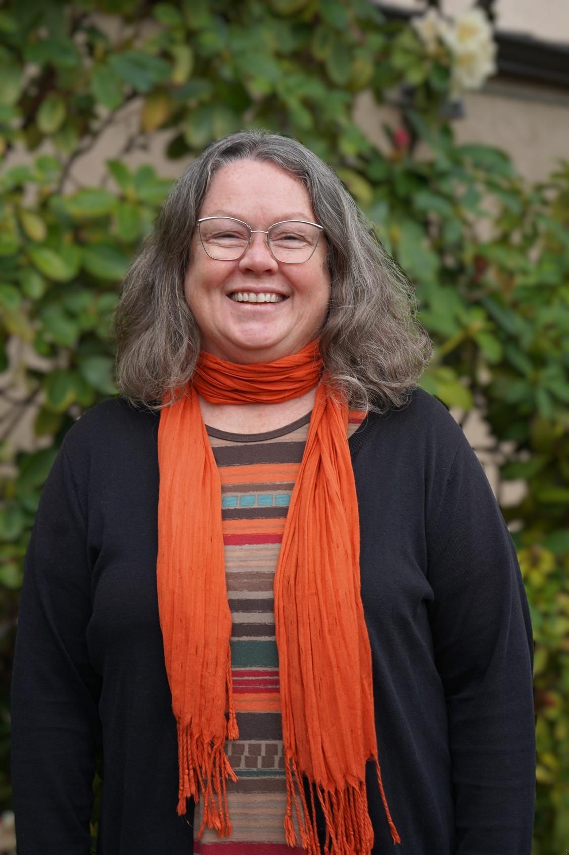 Woman smiling, wearing glasses, orange scarf, striped top, black cardigan, outdoors in front of greenery.