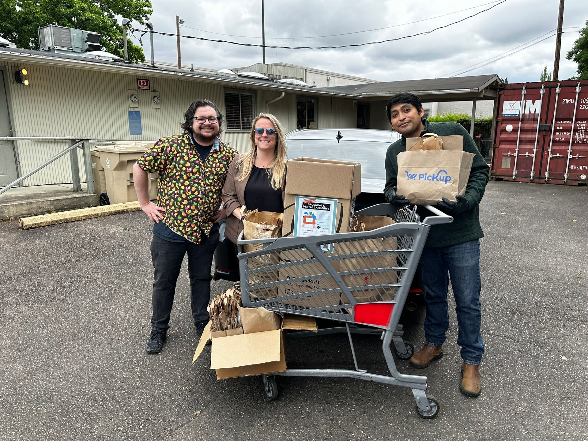 Three people smile in front of a car with a cart full of donation items in front of them. On the left is a short man in a colorful button-down shirt and black pants, with shoulder-length wavy hair, a beard, and glasses. In the middle is a short woman with blond hair wearing sunglasses, a brown cardigan over a black shirt. On the left is a taller man with short dark hair, wearing a black jacket, blue jeans, and tan boots, holding a bag of donation items. 