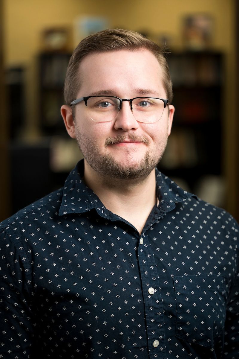 Man with glasses, button-down shirt, looking directly at the camera with a slight smile; indoors, blurred background.