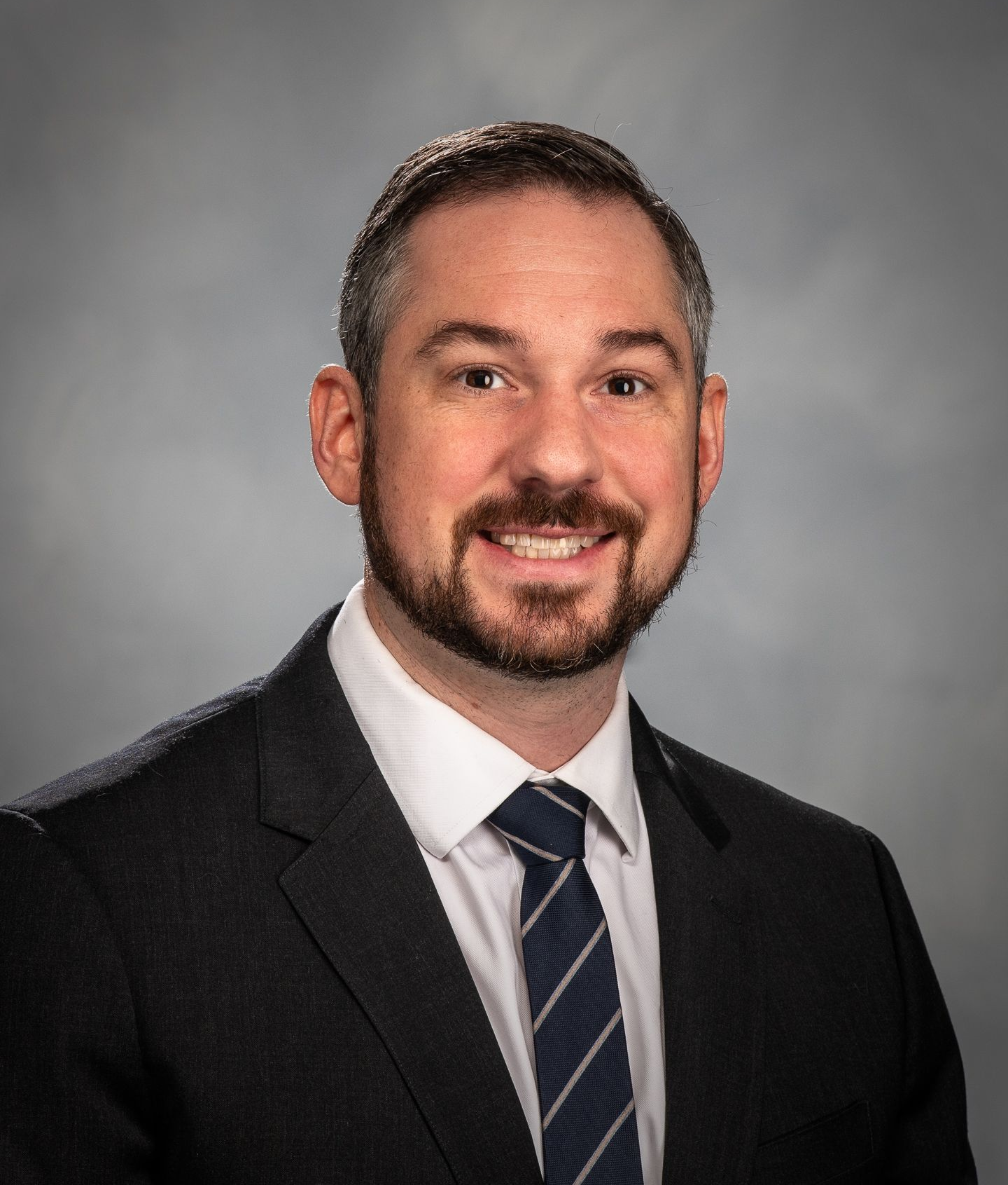 Man in suit smiles at the camera, dark hair and beard, blue tie, studio background.