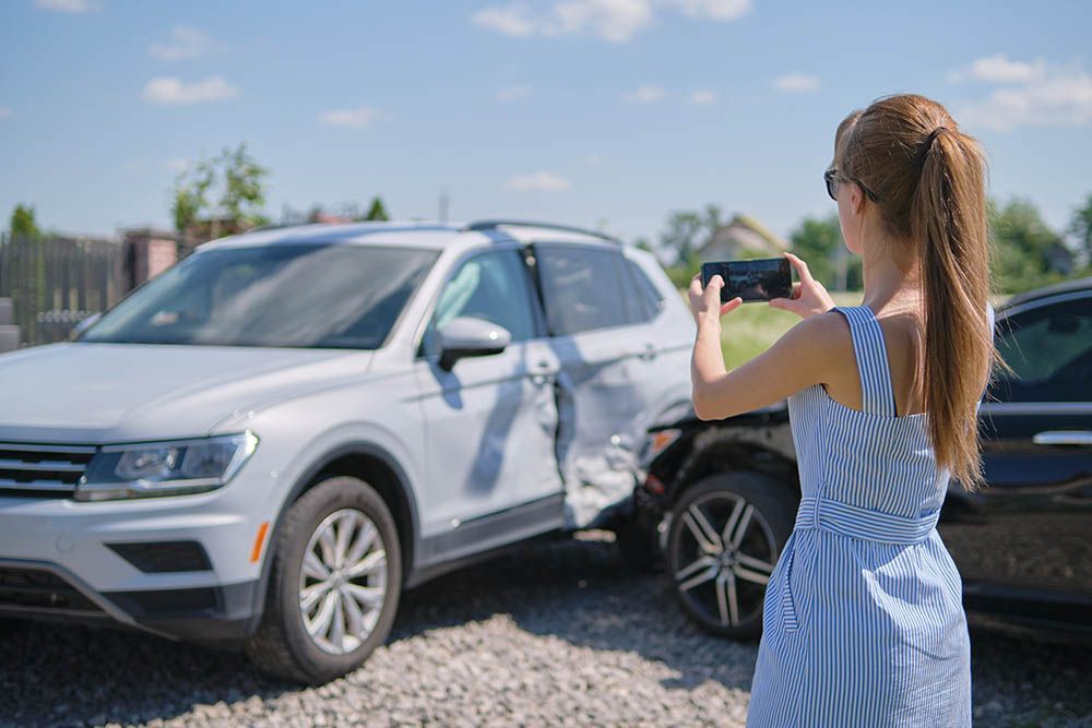 Woman in a striped dress photographs car damage with a phone after a car accident outdoors.