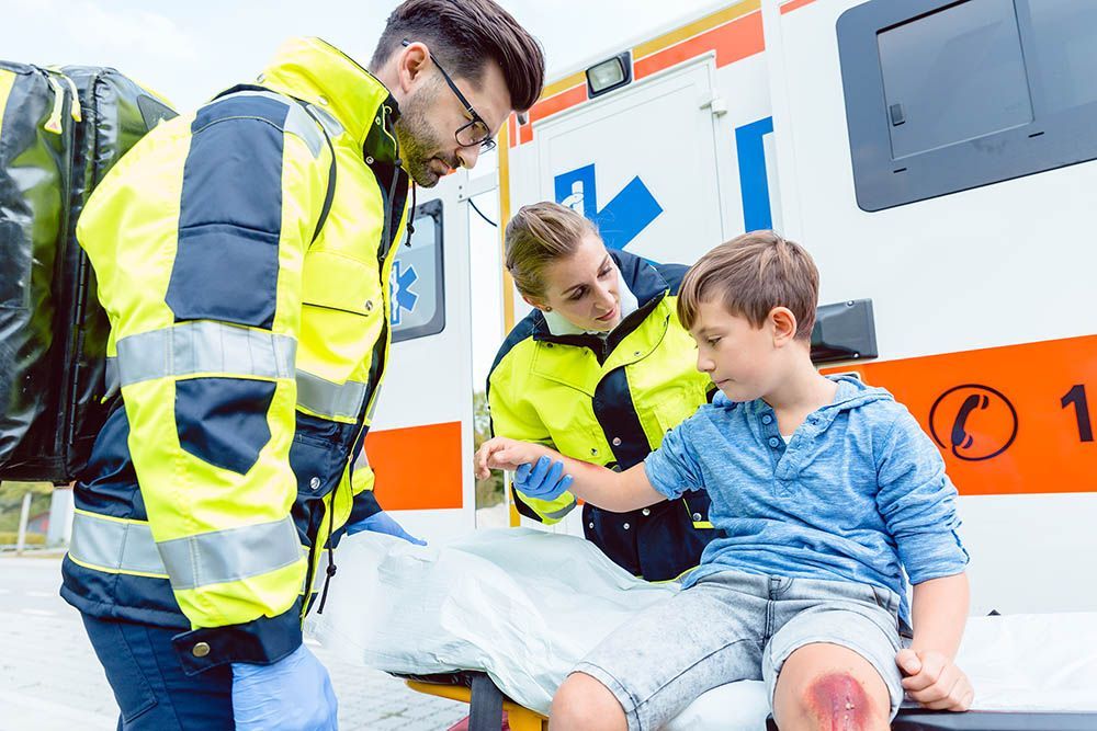 Paramedics helping a young boy with a scraped knee. An ambulance is in the background.