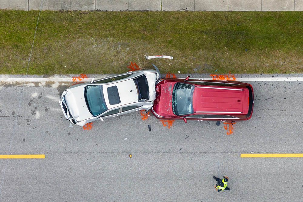 Overhead view of a car accident: silver sedan and red SUV crashed on a road, marked with orange cones.