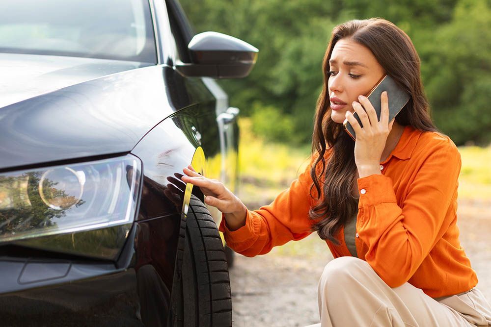 Woman with car damage on phone, looking distressed; outdoors.