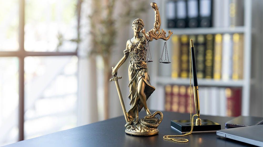 Statue of Lady Justice holding scales and sword on a desk in an office, with bookshelves visible.
