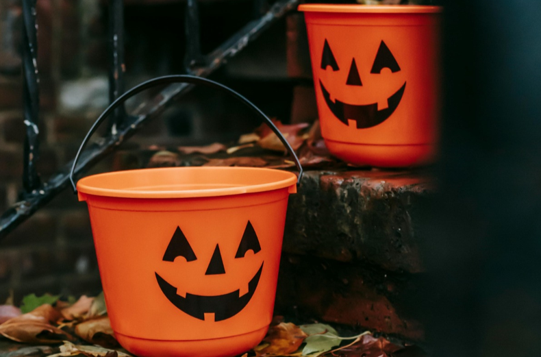 Two orange Halloween candy buckets with a jack'o'lantern smile on a outdoor stone steps, one is a step above the other. Fall leaves strew the steps, and a black metal rail is visible in the background.